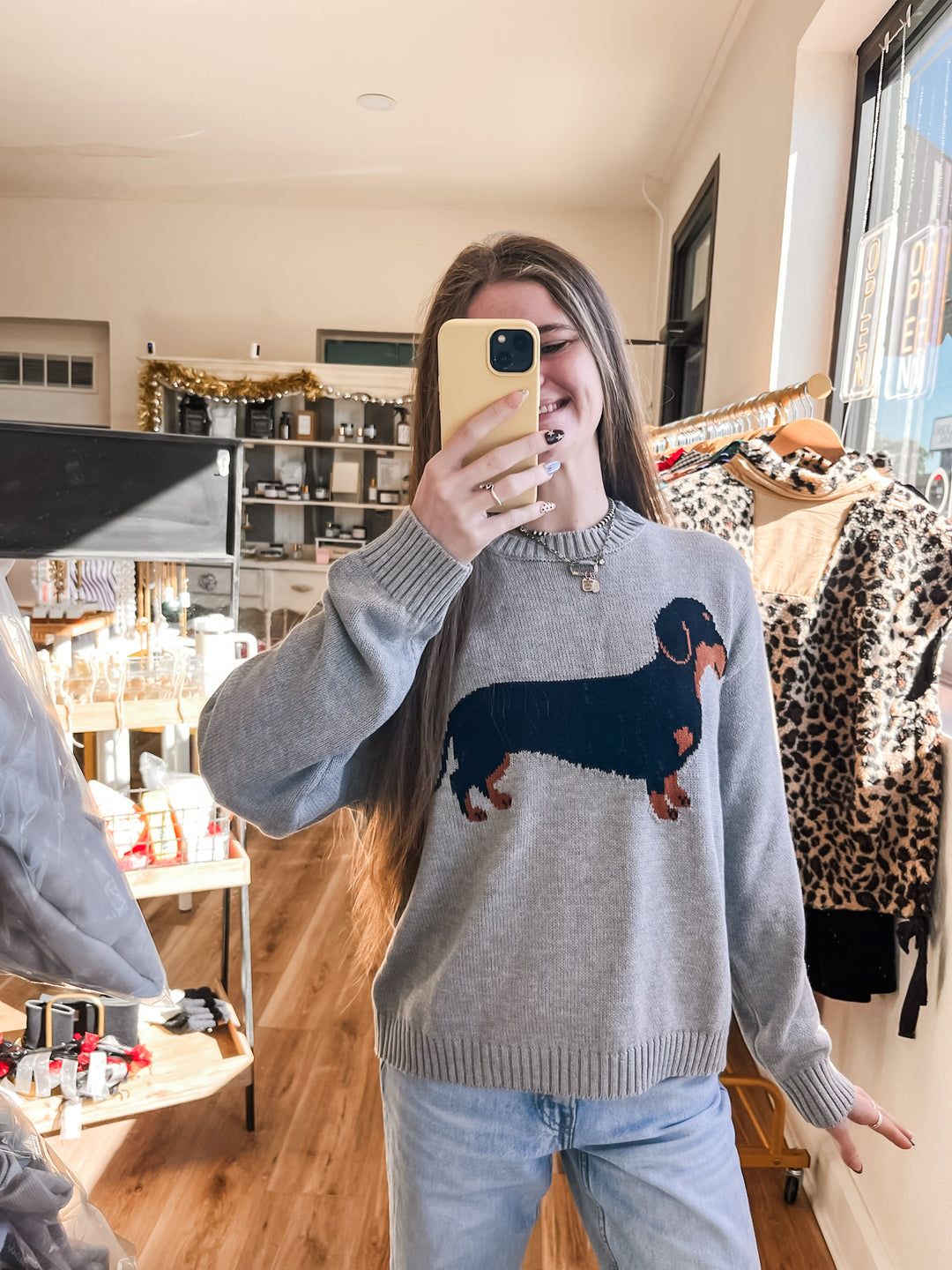 A woman takes a mirror selfie in a clothing boutique wearing a light gray knit sweater with a large black and tan dachshund graphic on the front, paired with light blue jeans.