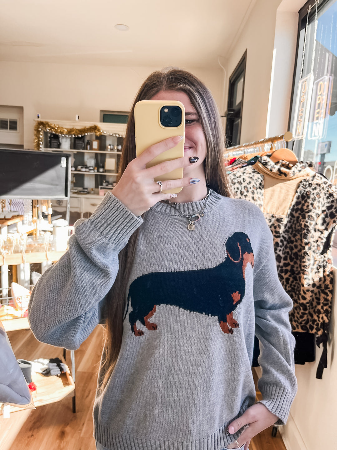 A woman takes a mirror selfie in a clothing boutique wearing a gray knit sweater with a large black and brown dachshund graphic on the front.