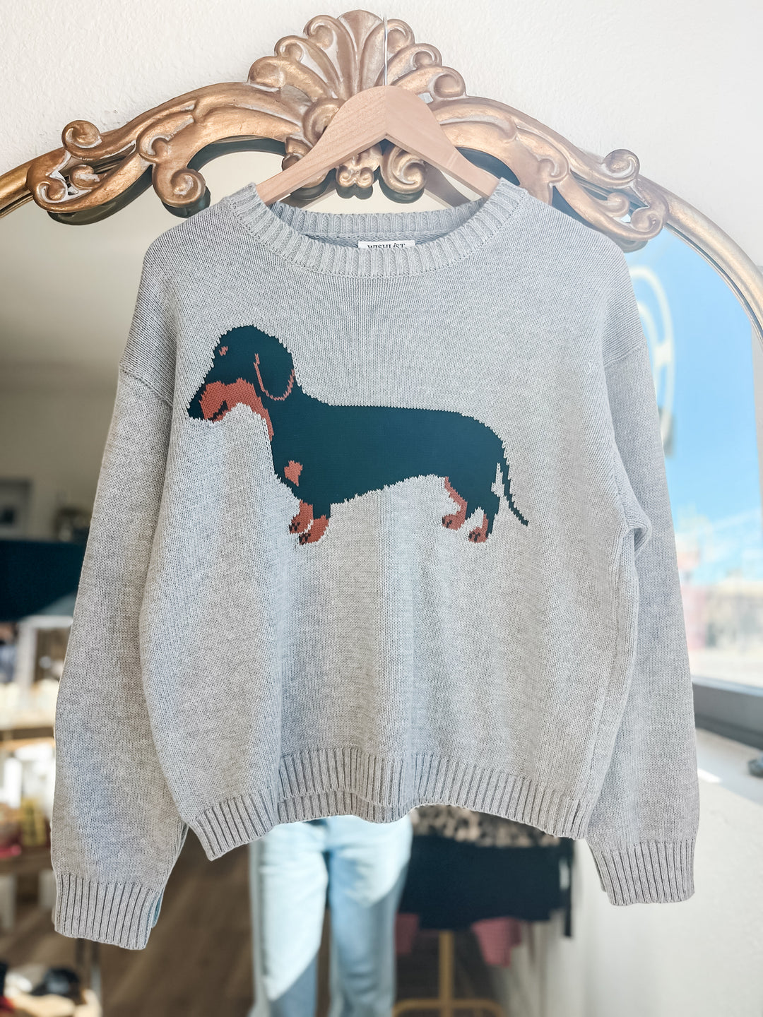 A light gray knit sweater hanging on a wooden hanger, featuring a large black and tan dachshund graphic across the chest. The sweater is displayed in front of an ornate gold mirror.
