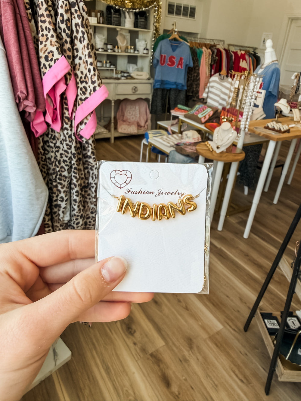 A hand holding a white jewelry card featuring gold-colored enamel lettering that reads 'INDIANS' with a small heart logo and 'Fashion Jewelry' above it, set against a blurred boutique background.