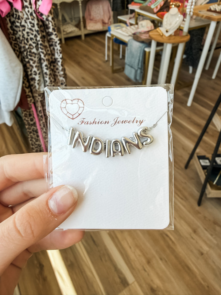 A hand holding a clear plastic-wrapped jewelry card featuring a silver-toned necklace with the word "INDIANS" in raised lettering, displayed in a retail store setting.