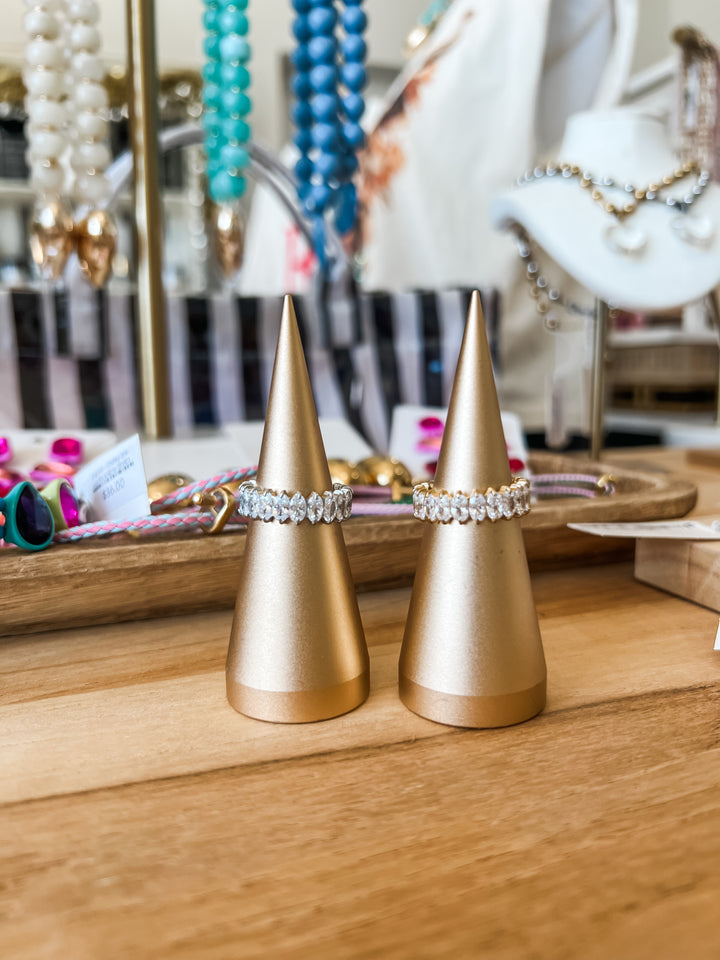 Two gold cone ring holders displaying a silver band and a gold band with clear rhinestones, set on a wooden counter with a blurred jewelry store background.