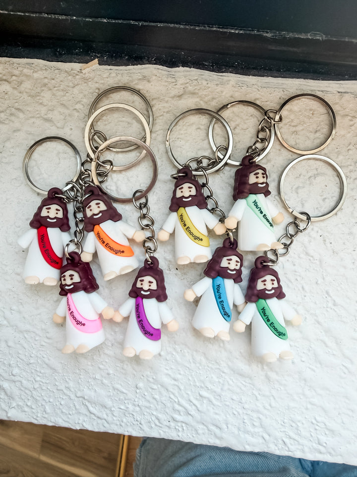 Six small Jesus keychains with colorful sashes and brown hair are arranged on a textured white surface. Each keychain has silver metal key rings and a chain.