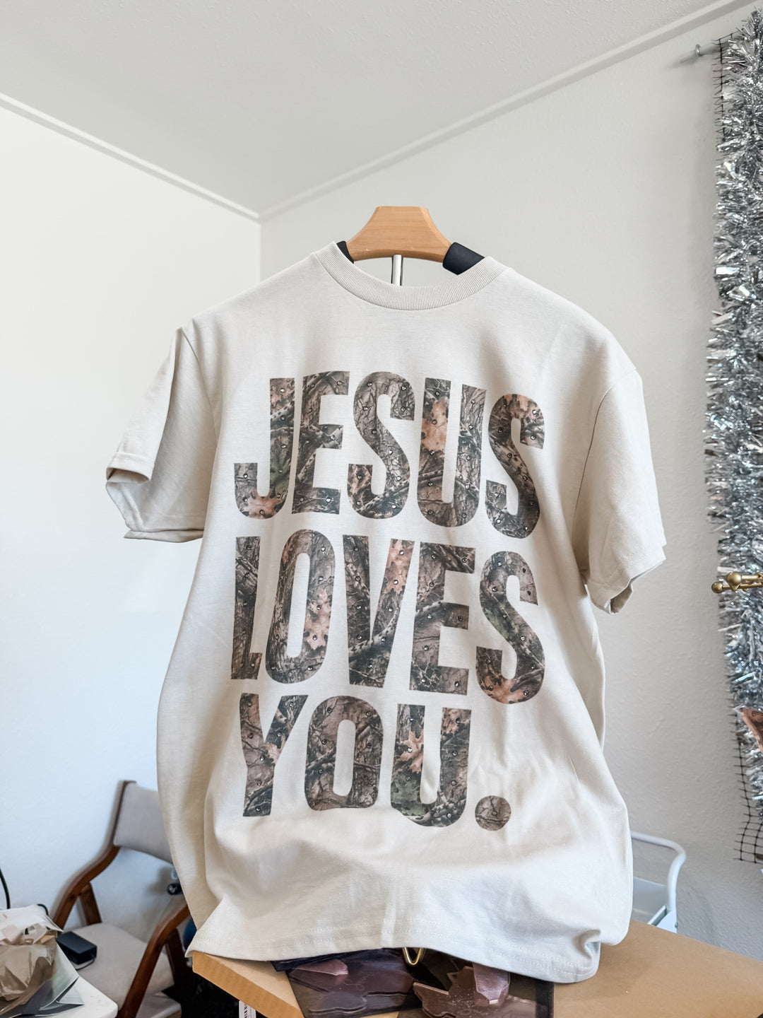 A cream-colored short-sleeve t-shirt hanging on a wooden hanger, featuring the text 'JESUS LOVES YOU.' in a distressed camouflage pattern.