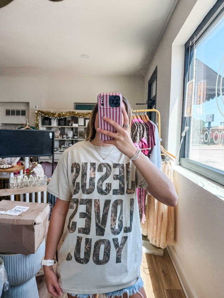 A person taking a mirror selfie in a boutique, wearing a light beige graphic t-shirt with the words &