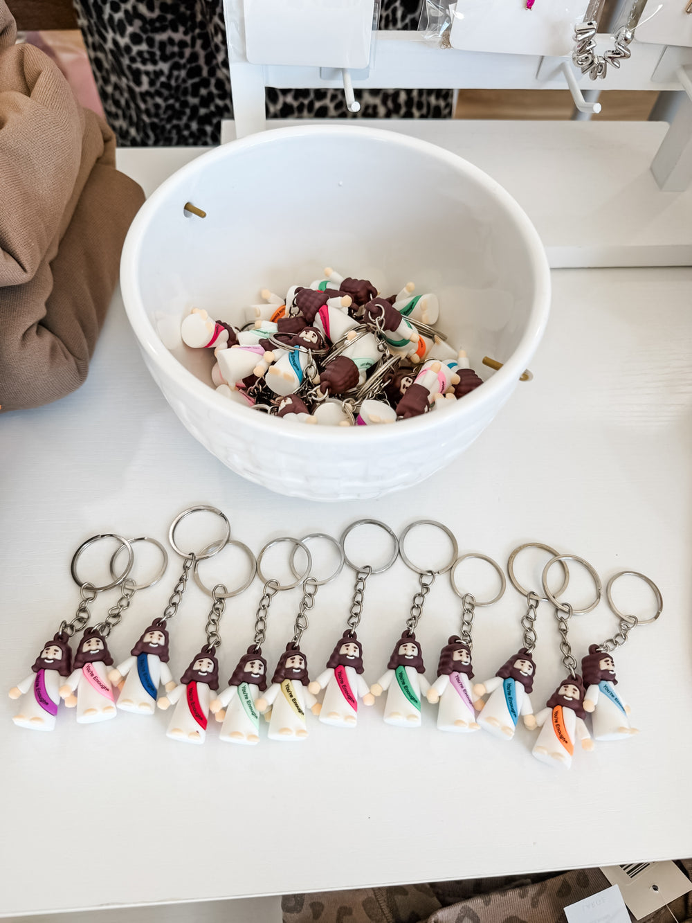 A collection of small keychains shaped like Jesus in white outfits, displayed on a white table with a white bowl filled with similar keychains behind them.
