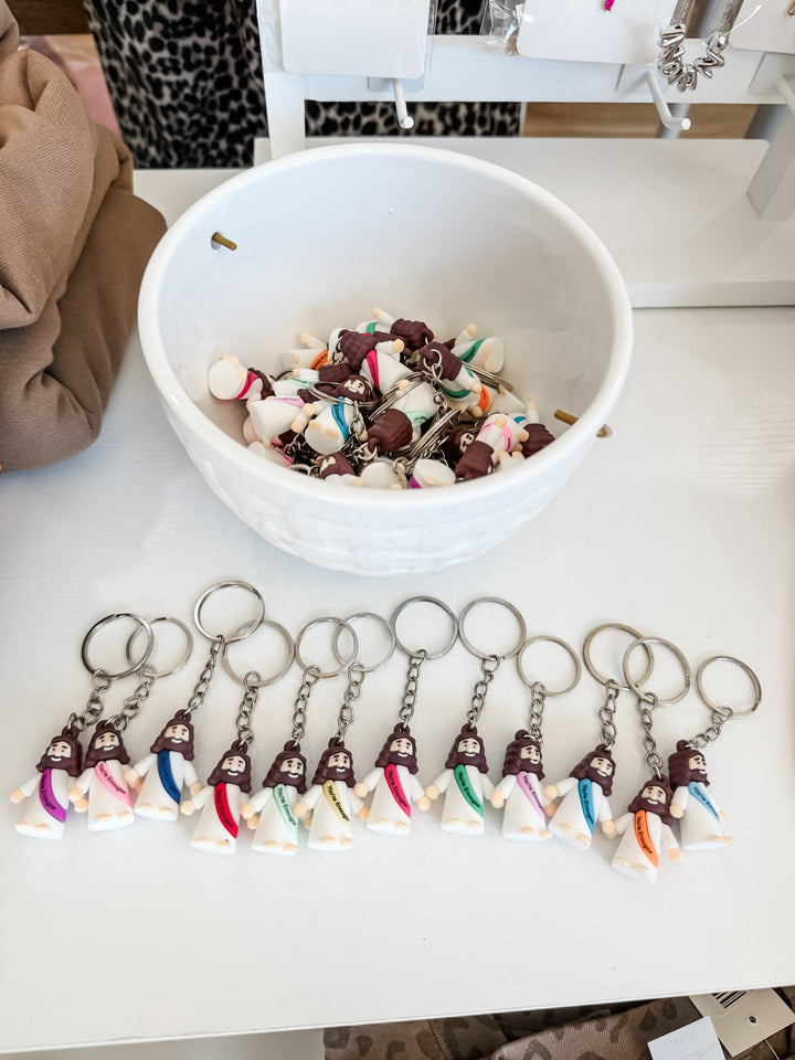 A collection of small keychains shaped like Jesus in white outfits, displayed on a white table with a white bowl filled with similar keychains behind them.