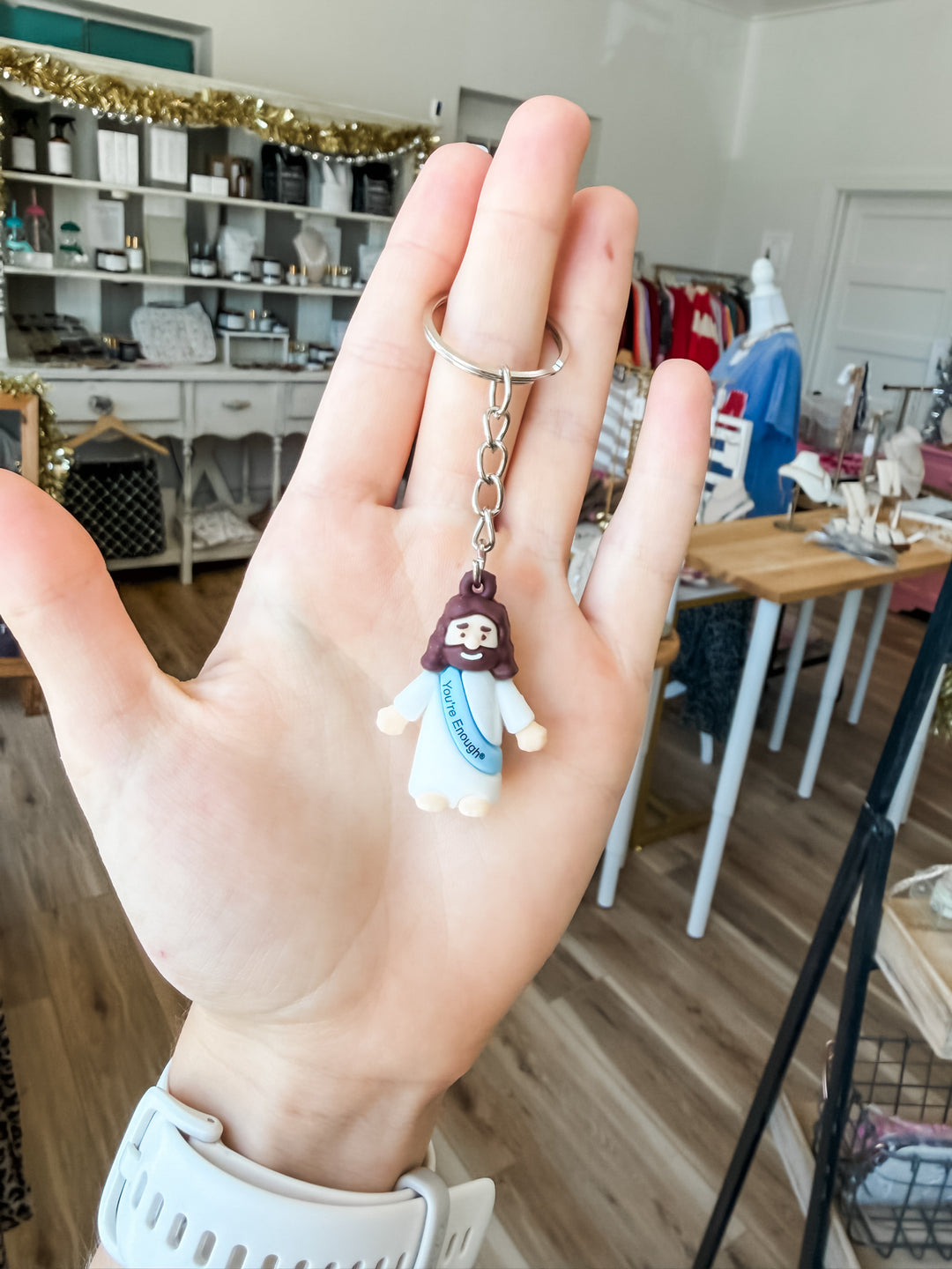 A hand holding a small Jesus keychain charm with a silver split ring, set against a blurred boutique background.