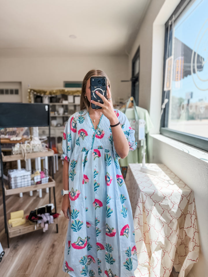 A woman takes a mirror selfie in a boutique wearing a light blue kaftan dress with a pink flower and green leaf pattern.