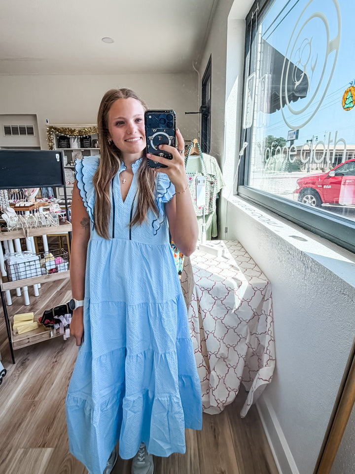 A woman takes a mirror selfie in a boutique while wearing a light blue tiered midi dress with ruffled sleeves and a high collar. The shop interior is visible behind her, with clothing racks, shelves, and a window.