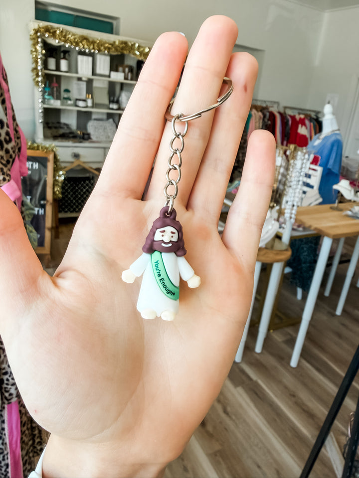 A hand holding a small Jesus keychain charm with a silver split ring, set against a blurred boutique background.