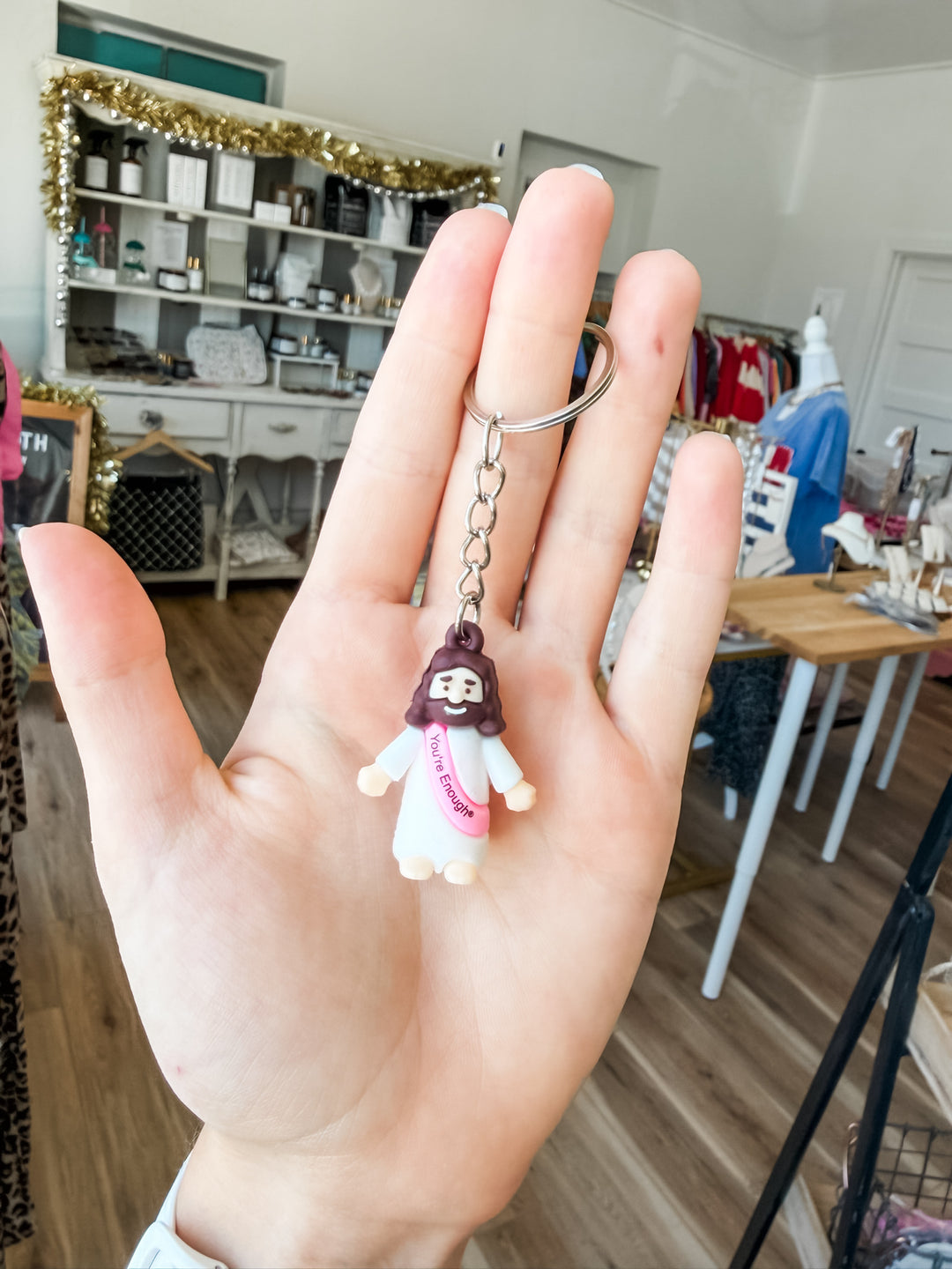 A hand holding a small Jesus keychain charm in front of a blurred boutique background.