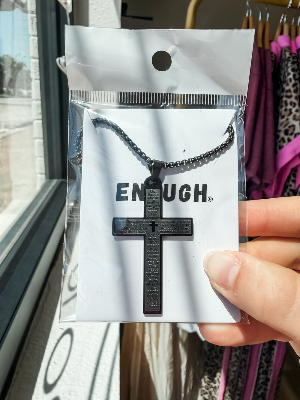 A black cross pendant necklace with engraved text, displayed on a white card with the brand name 'ENOUGH' and held by a hand inside a store.