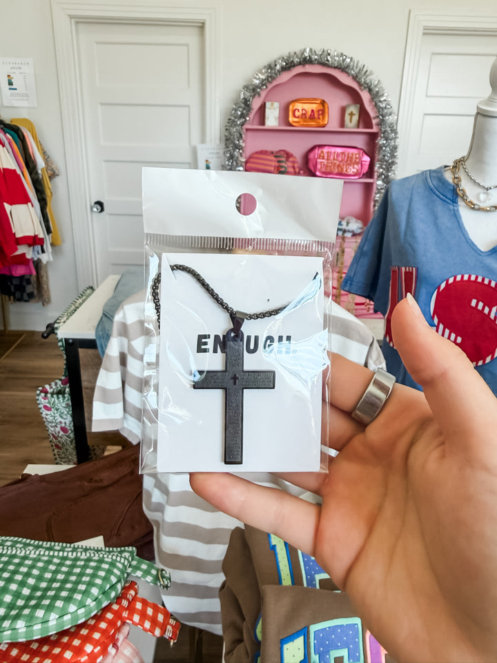 A hand holding a black cross pendant necklace on a white card with the word &