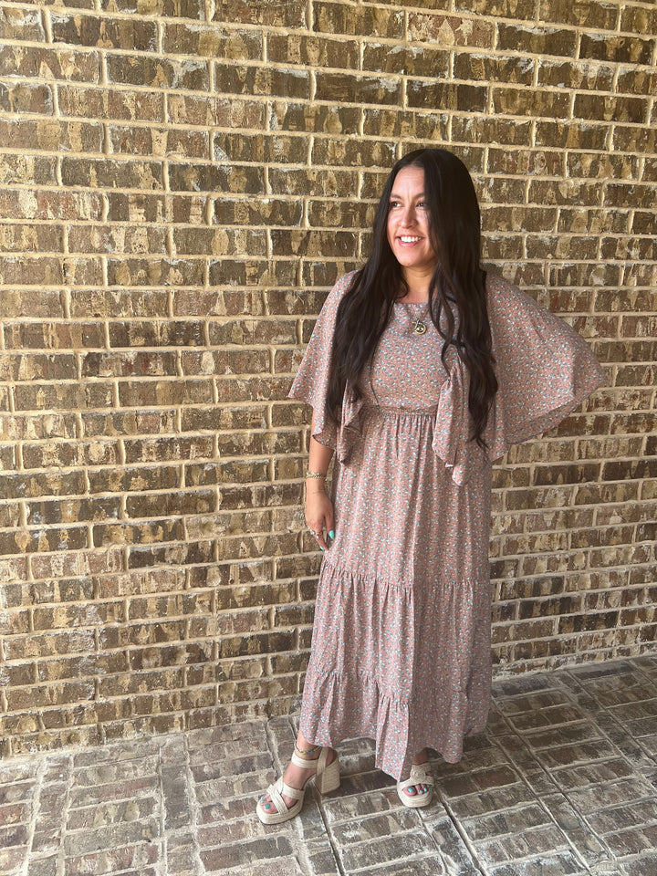 A woman with long dark hair stands against a tan brick wall wearing a mauve tiered maxi dress with a small floral pattern and matching sheer flutter sleeves, paired with beige heeled sandals.