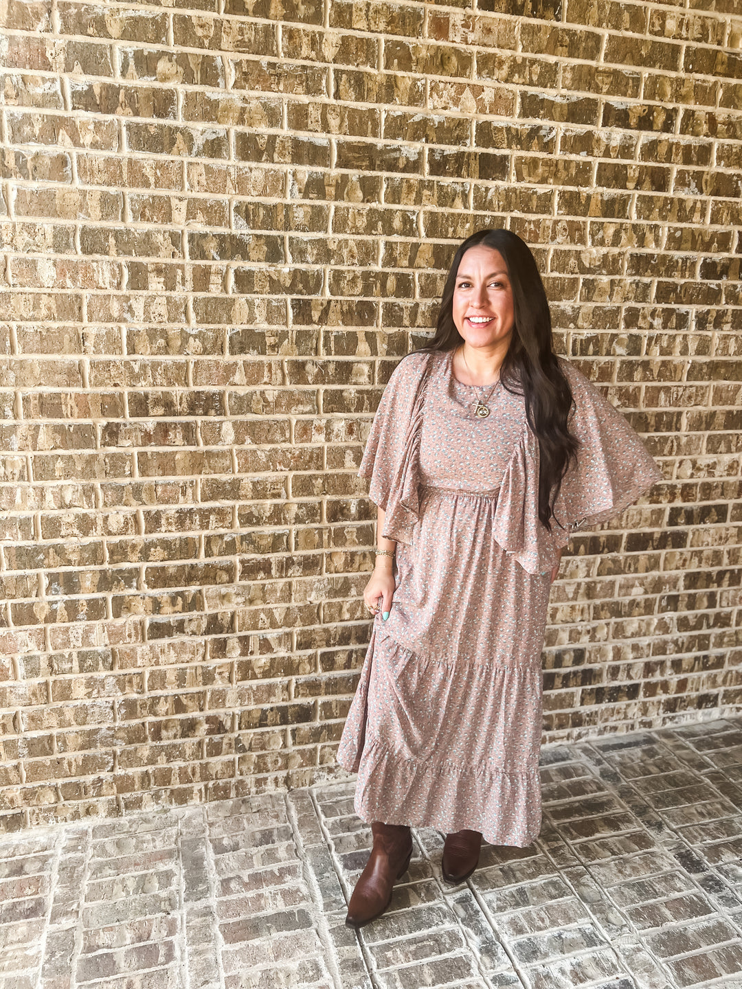 A woman wearing a dusty pink tiered maxi dress with flutter sleeves and brown ankle boots, standing in front of a tan brick wall.