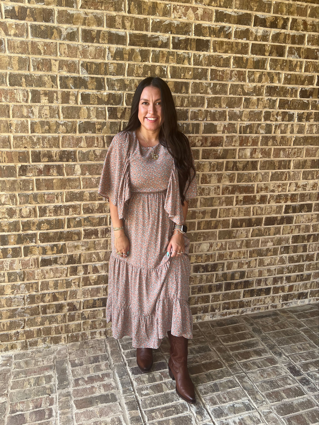 A woman stands in front of a tan brick wall wearing a mauve  flutter-sleeve, tiered midi paired with brown knee-high boots.