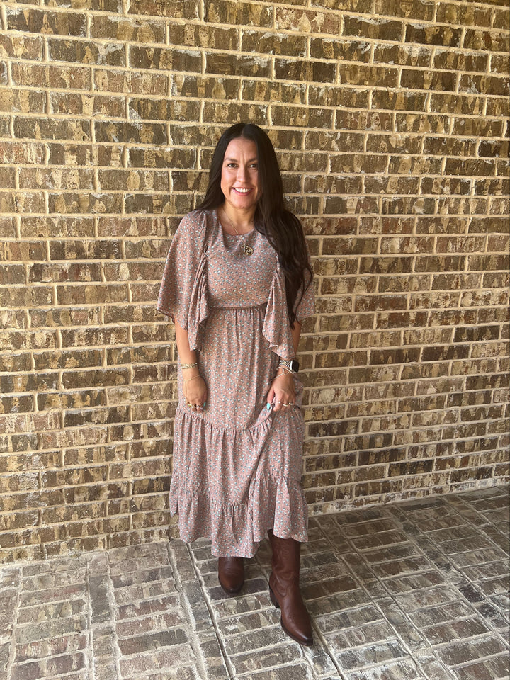 A woman stands in front of a tan brick wall wearing a mauve  flutter-sleeve, tiered midi paired with brown knee-high boots.