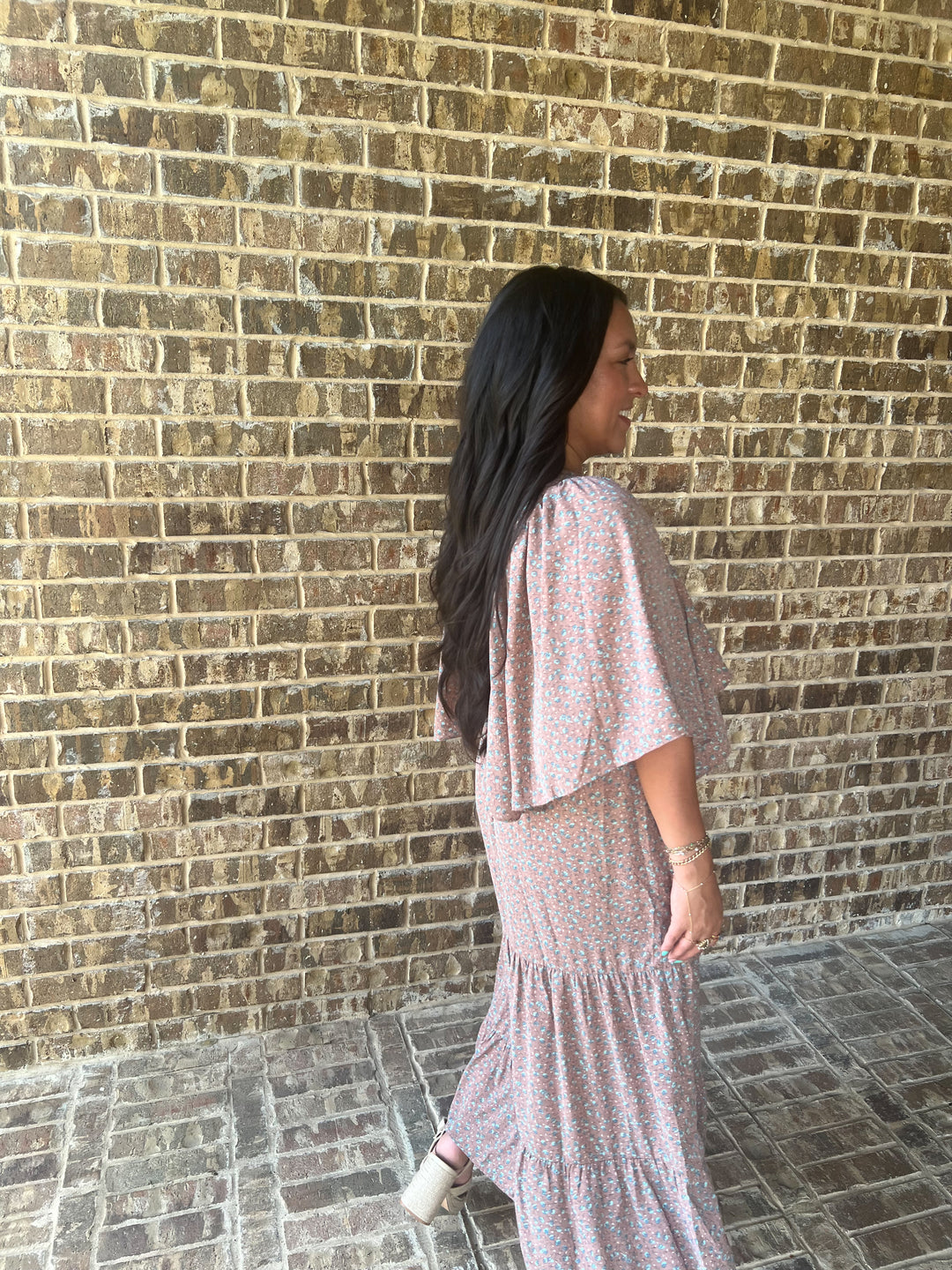A woman with long dark hair is shown from the back and side, wearing a mauve tiered maxi dress with flutter sleeves, standing against a brown brick wall.