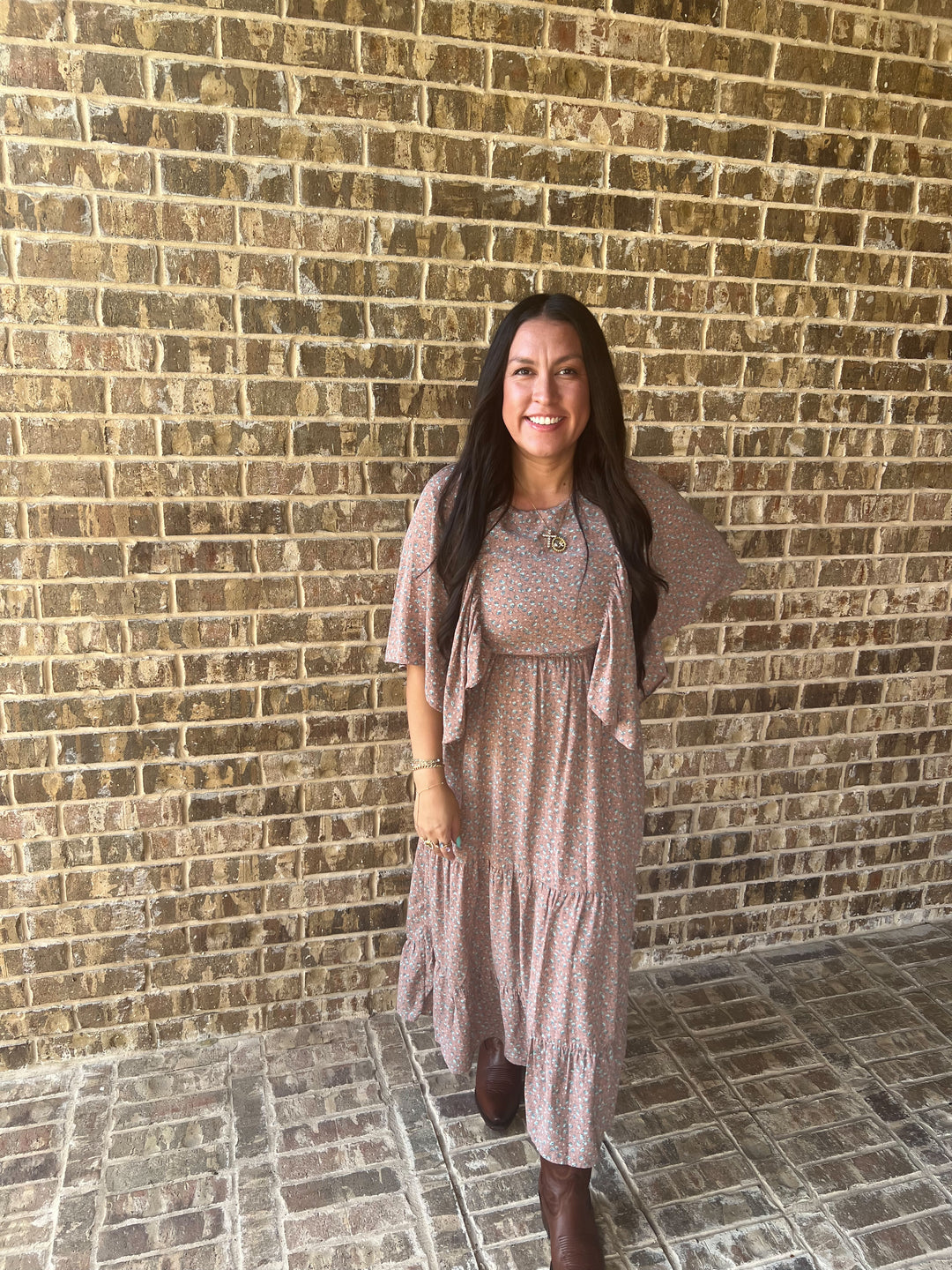 A woman with long dark hair stands smiling in front of a tan brick wall, wearing a loose, short-sleeve tiered maxi dress in a muted mauve-brown color with a small light pattern, paired with brown boots.