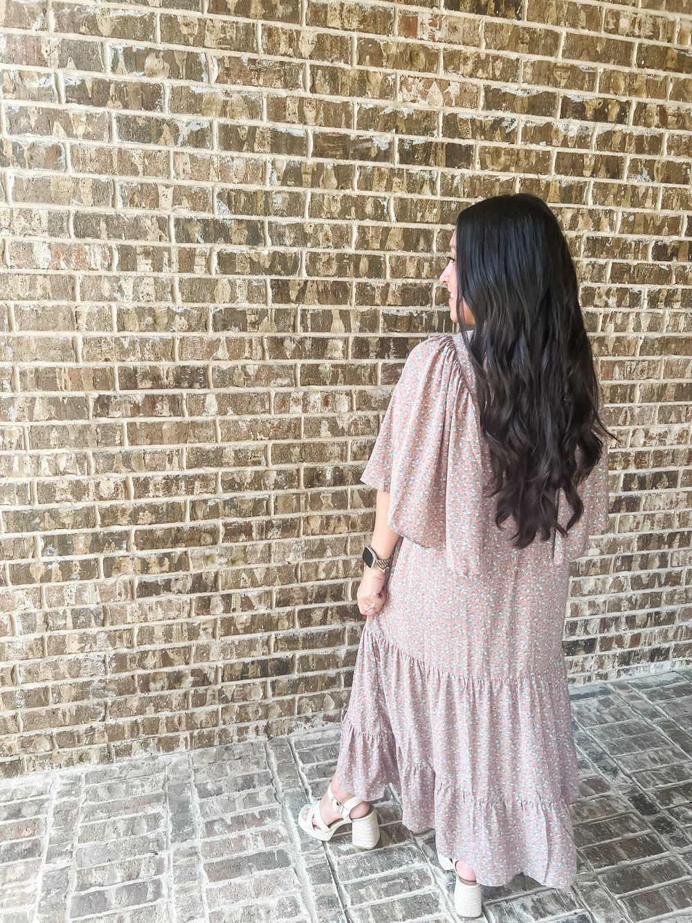 A woman with long dark hair is seen from behind, wearing a mauve tiered maxi dress and white block-heel sandals, standing against a brown brick wall.