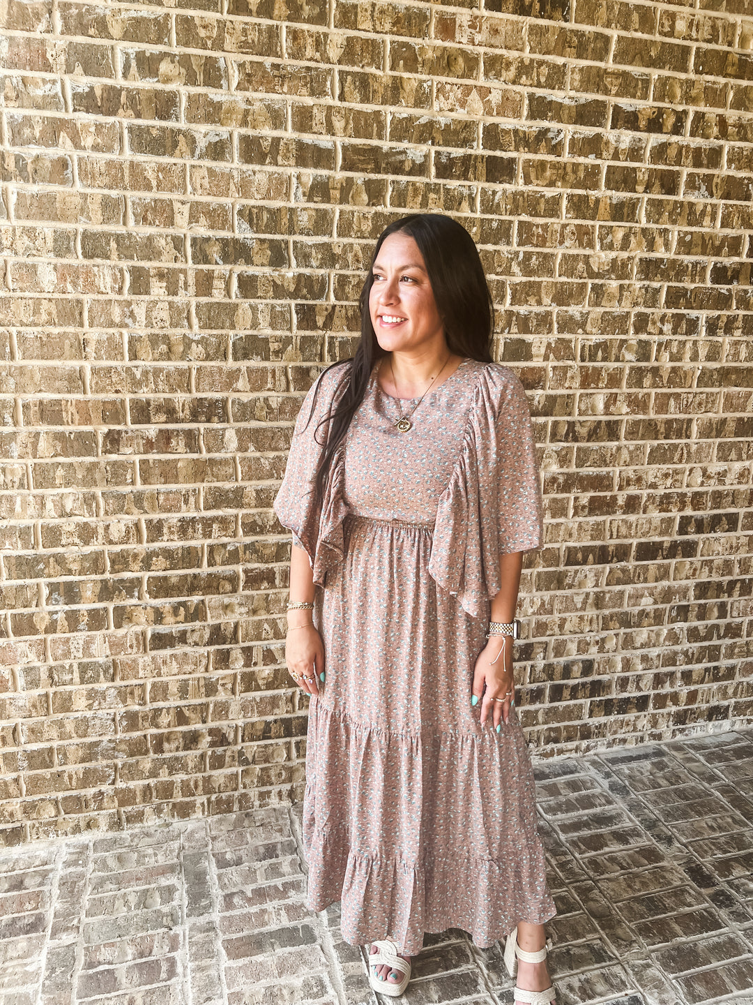 A woman wearing a tiered midi dress with a small floral print in dusty pink and beige, standing in front of a brick wall.