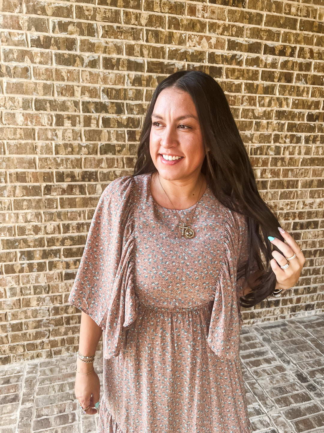 A woman with long dark hair stands in front of a brick wall wearing a loose, short-sleeve floral dress in muted pink, beige, and blue tones. She is smiling and holding her hair.