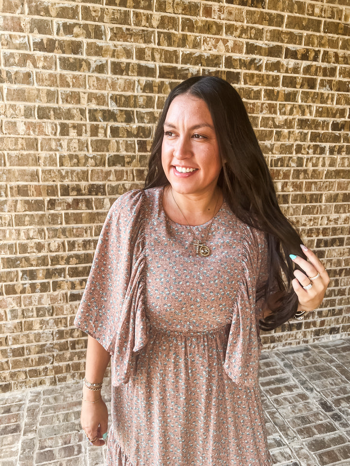 A woman with long dark hair stands in front of a brick wall wearing a loose, short-sleeve floral dress in muted pink, beige, and blue tones. She is smiling and holding her hair.