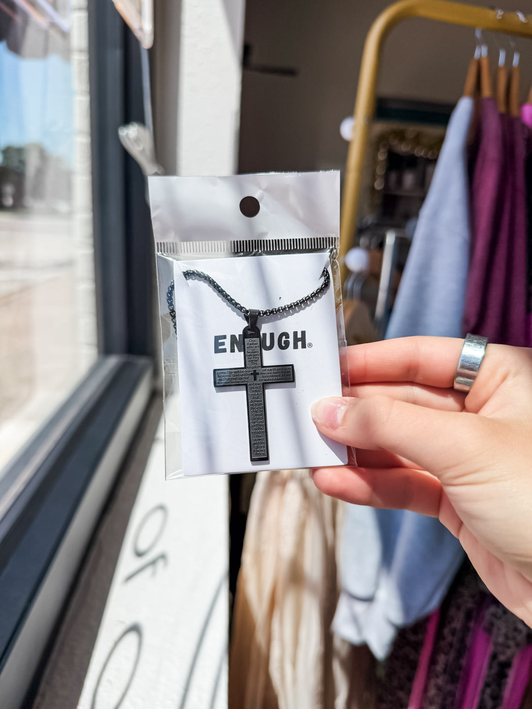 A hand holds a black cross pendant on a matching chain, packaged on a white card printed with the word 'ENOUGH.', in a retail store setting with clothing racks blurred in the background.