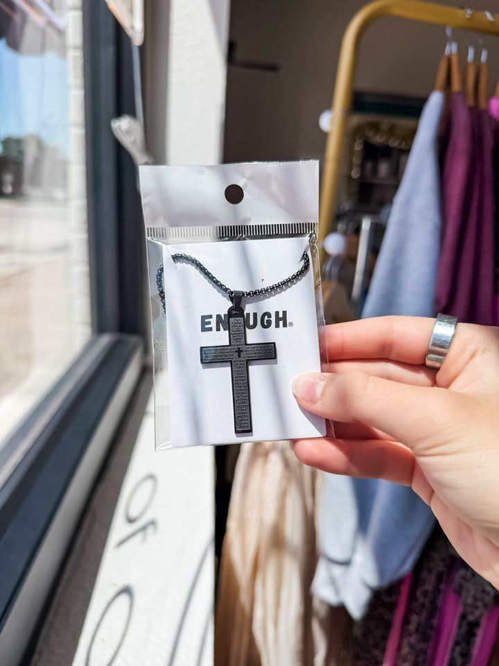 A hand holds a black cross pendant on a matching chain, packaged on a white card printed with the word &