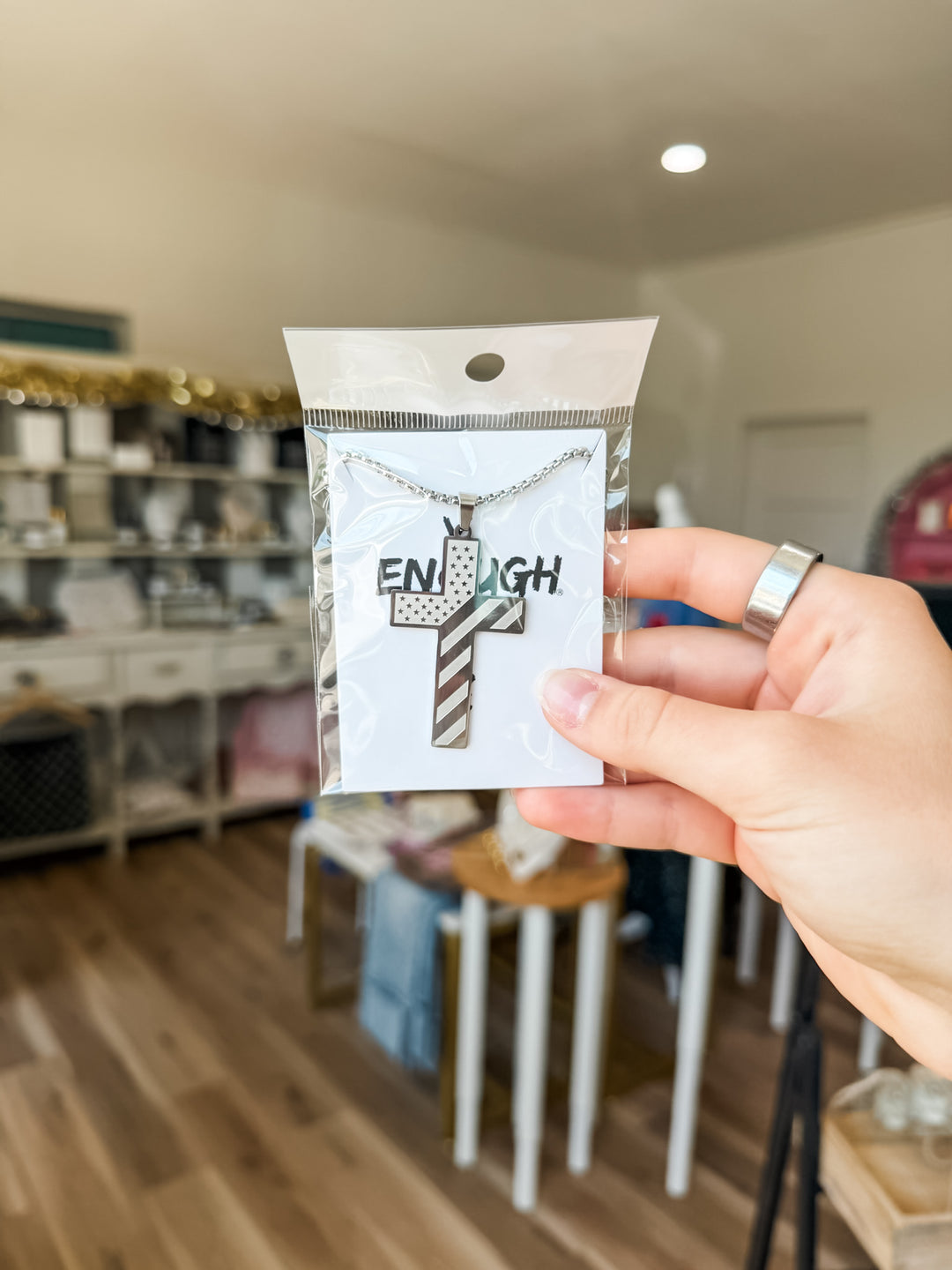 A hand holding a silver cross pendant necklace on a white backing card that reads 'ENOUGH'. The background shows a blurred boutique interior with wooden floors and shelves.