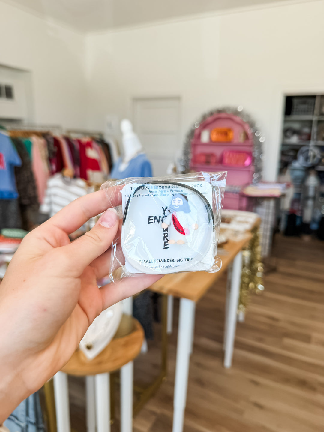 A hand holds a small packaged round item in front of a blurred boutique interior. The package contains a black and white bracelet  and mini Jesus figurine that say "You&
