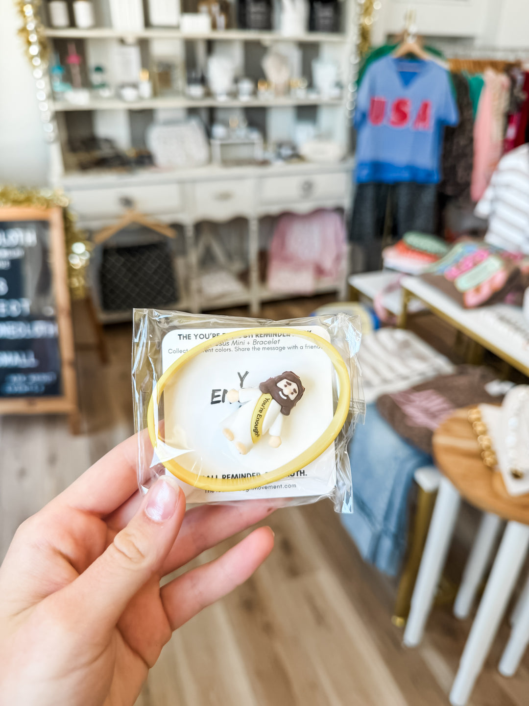 A hand holding a packaged yellow bracelet with a small Jesus figurine in a retail store setting.