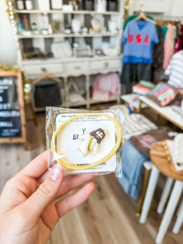 A hand holding a packaged yellow bracelet with a small Jesus figurine in a retail store setting.