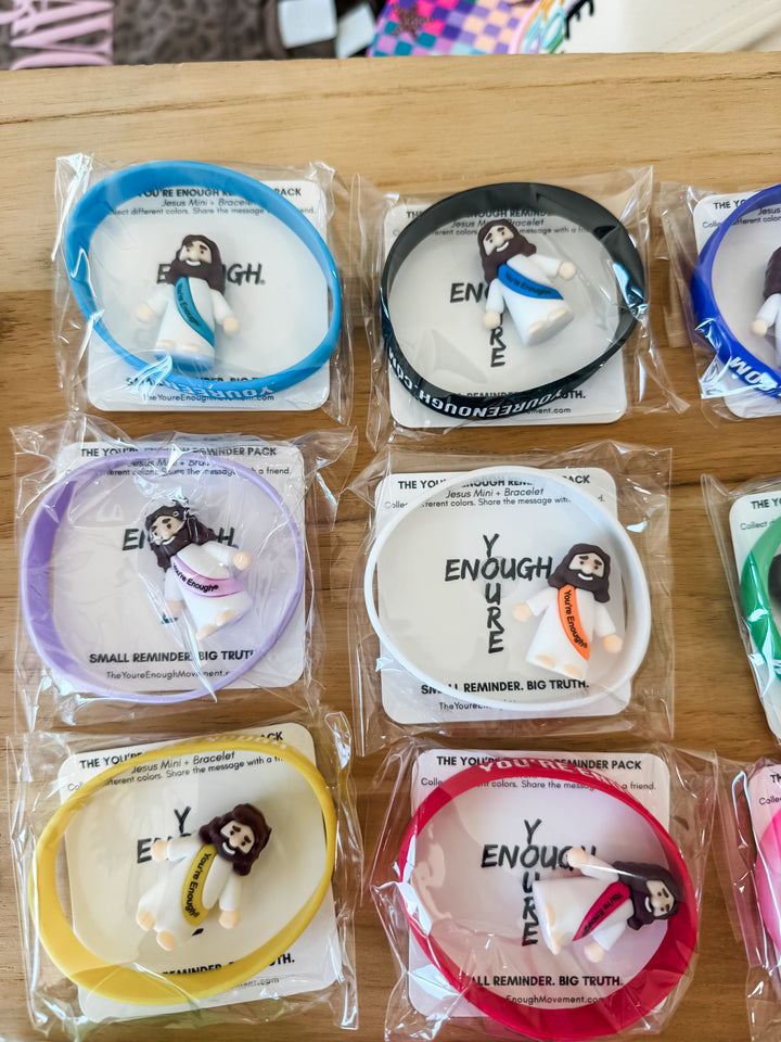 Several packaged bracelet + mini Jesus figurines arranged on a wooden surface, each with a Jesus figurine and printed backing card. The visible color options include blue, black, purple, yellow, red, and pink.