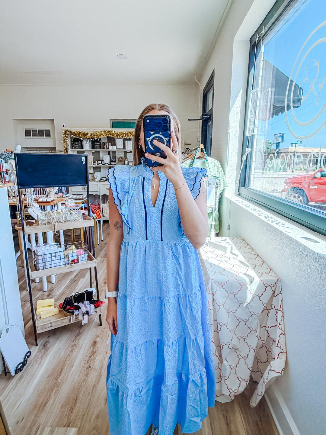 A woman takes a mirror selfie in a boutique wearing a light blue tiered maxi dress with ruffled sleeve details and a split neckline. The shop interior is visible in the background with clothing racks, shelves, and a window.
