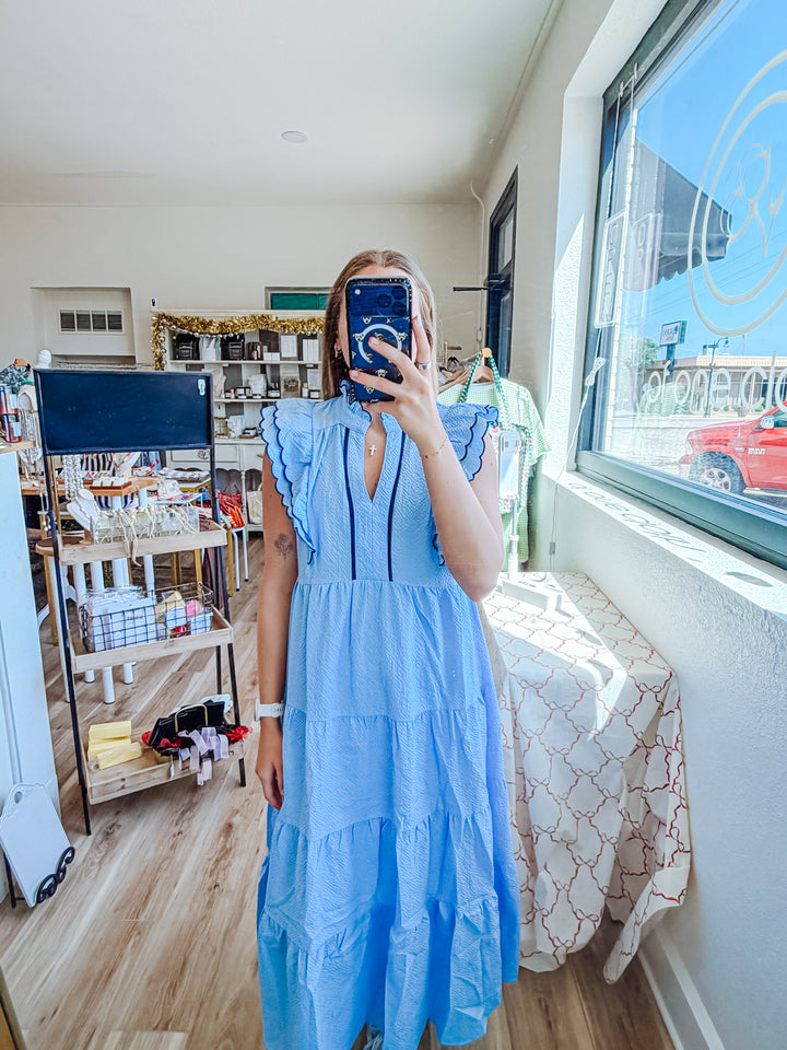 A woman takes a mirror selfie in a boutique wearing a light blue tiered maxi dress with ruffled sleeve details and a split neckline. The shop interior is visible in the background with clothing racks, shelves, and a window.