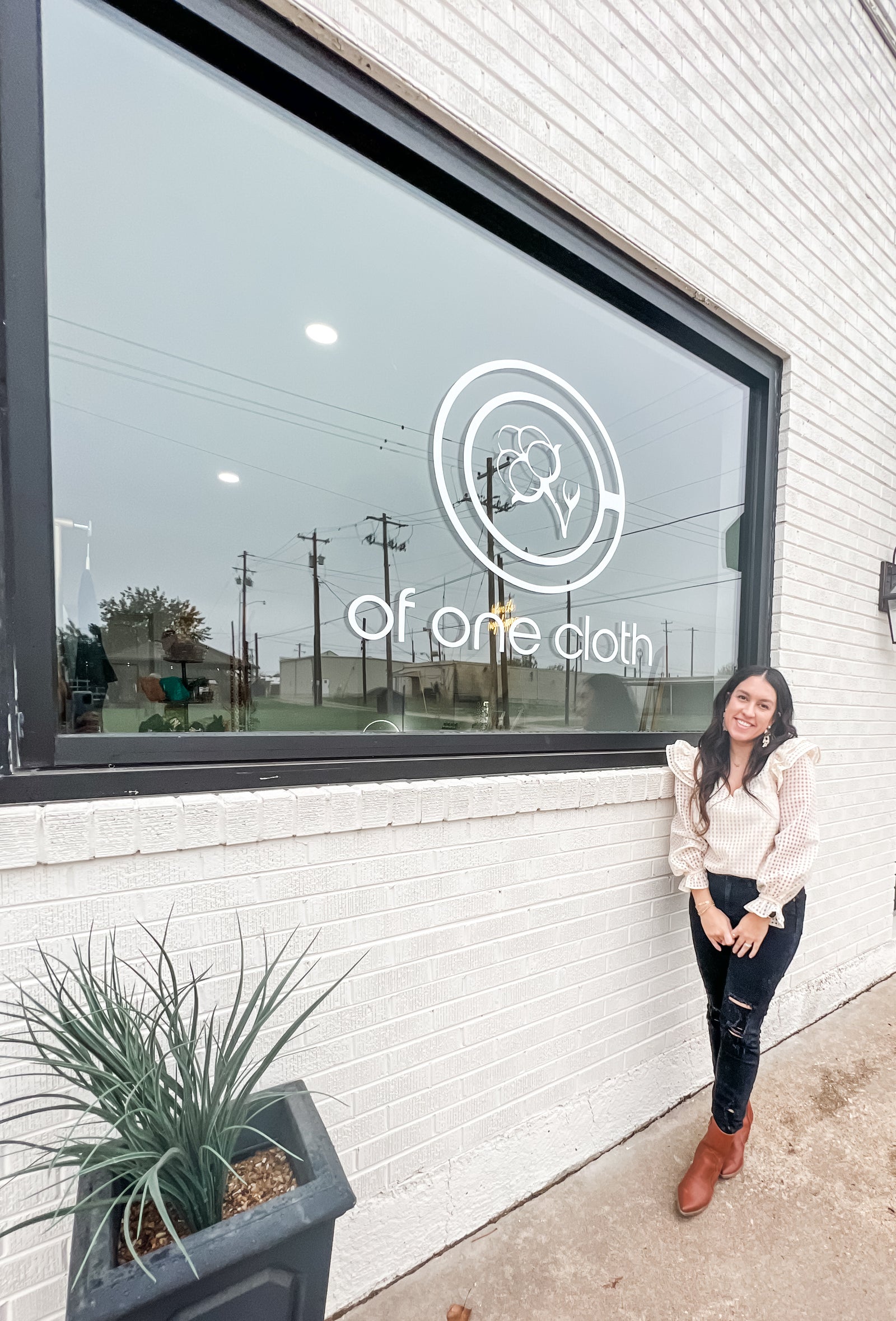 Woman in a white top and black jeans standing outside a store front with a window that has the "Of One Cloth" logo