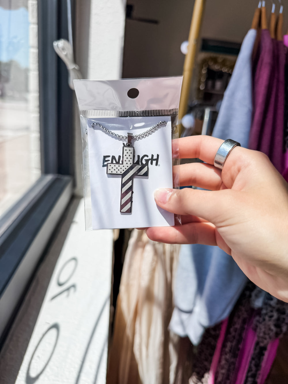 A hand holding a packaged necklace featuring a cross pendant with the word 'ENOUGH' and a striped barbershop tail design, with a clothing rack and window in the background.