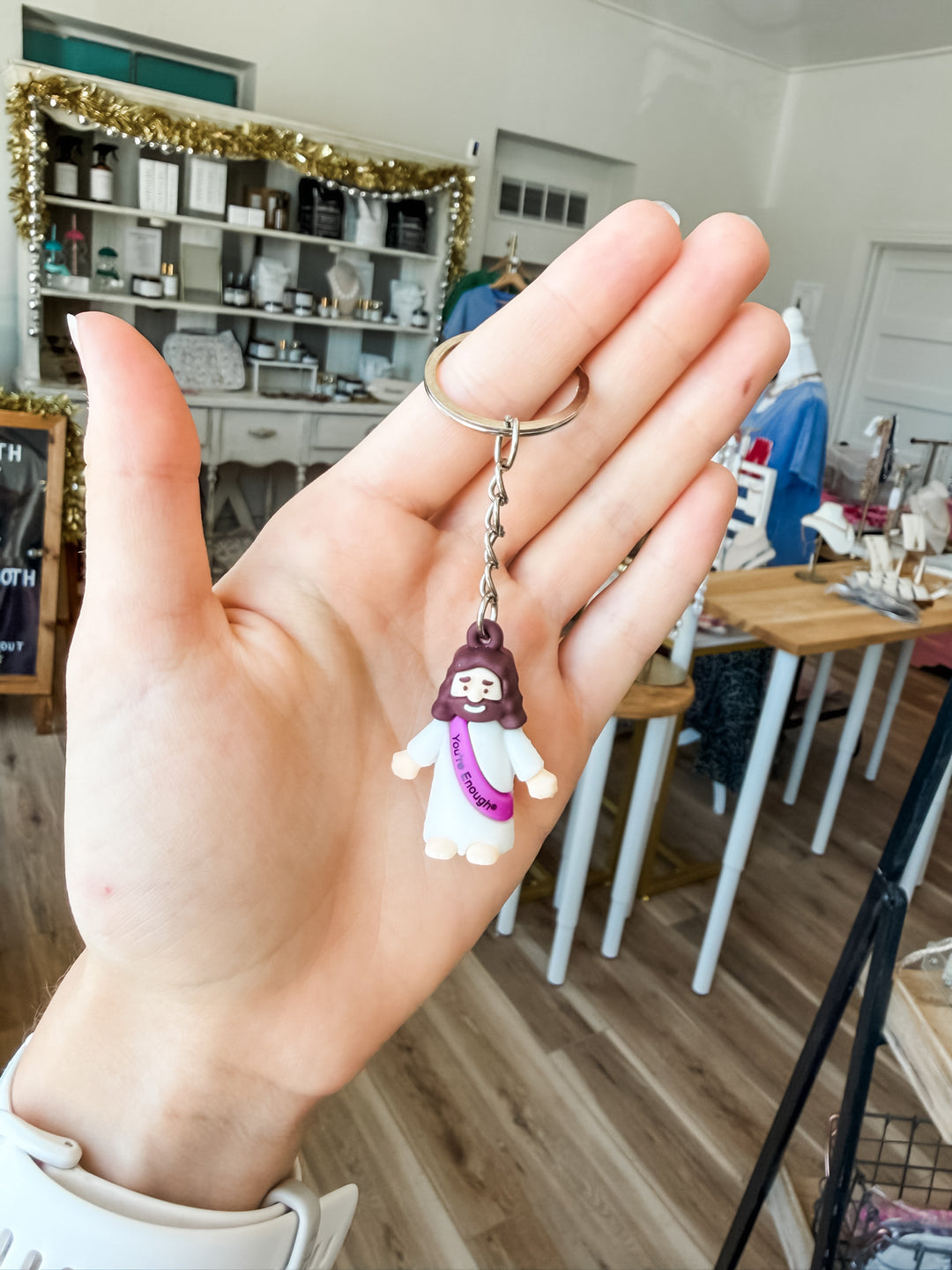 A hand holding a small Jesus keychain charm with a silver metal ring, set against a blurred boutique background.