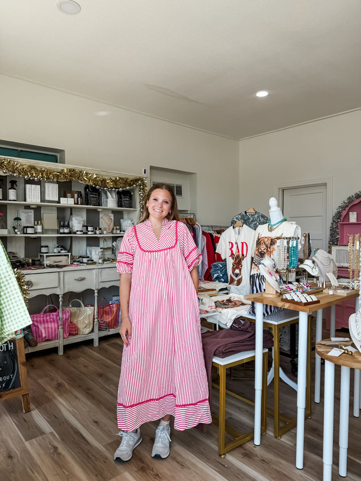 A woman wearing a pink and white vertically striped maxi dress with pink trim stands in a boutique interior. The shop has wood floors, white shelving with various items, and display tables with clothing and accessories.