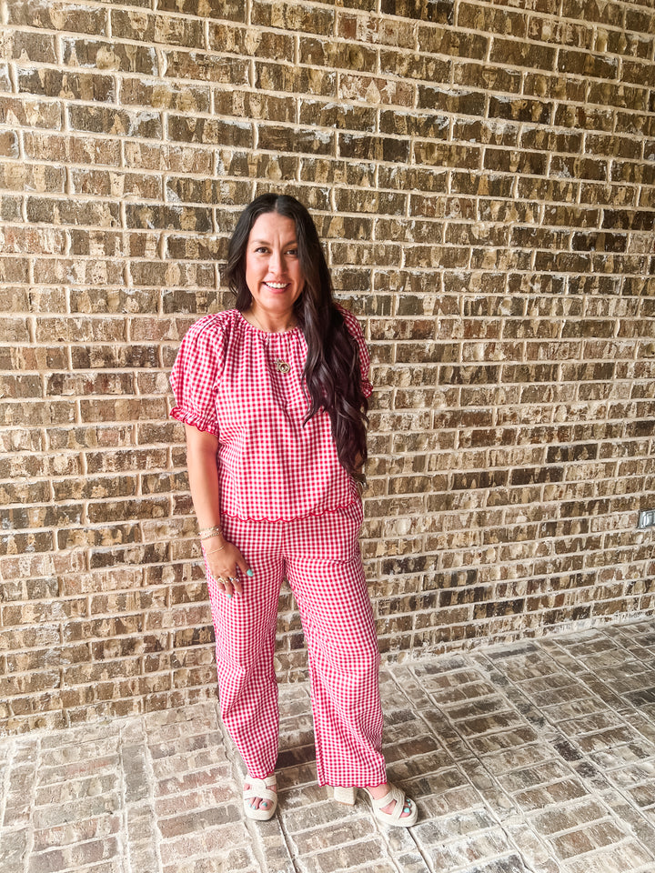 A woman wearing a red and white gingham two-piece set consisting of a short-sleeve top and matching wide-leg pants, standing in front of a brown brick wall.