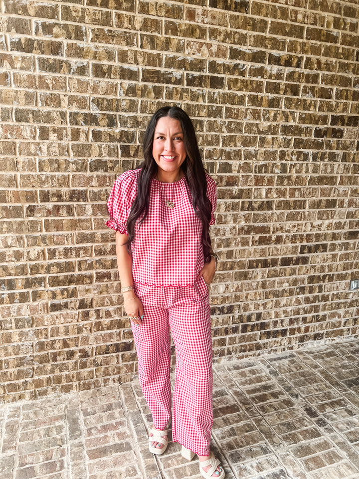 A woman wearing a red and white gingham two-piece set consisting of a short-sleeve top with puff sleeves and matching wide-leg pants, standing in front of a brown brick wall.