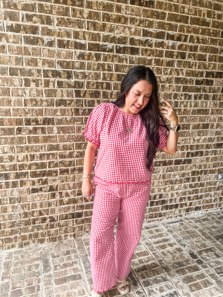 A woman wearing a red and white gingham two-piece set consisting of a short-sleeve top and matching wide-leg pants, standing against a brown brick wall.