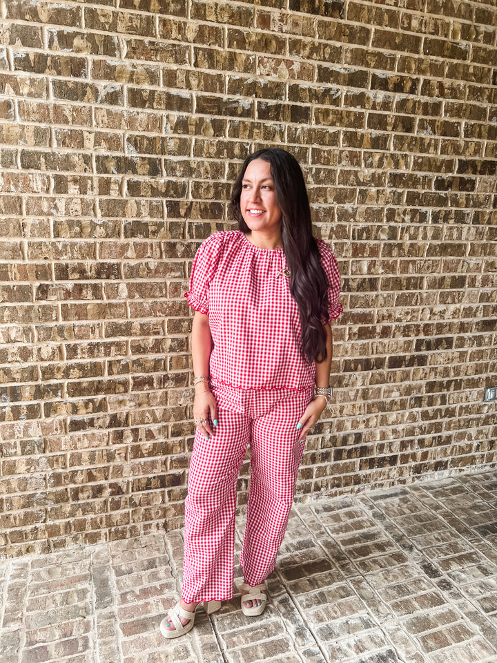 A woman wearing a red and white gingham two-piece set consisting of a short-sleeve top with puff sleeves and matching wide-leg pants, standing in front of a brown brick wall.