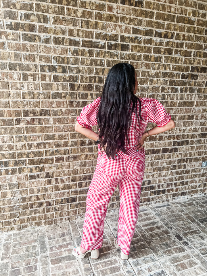 A woman with long dark hair stands with her back to the camera, wearing a pink and white gingham short-sleeve top and matching wide-leg pants, posing in front of a brick wall.