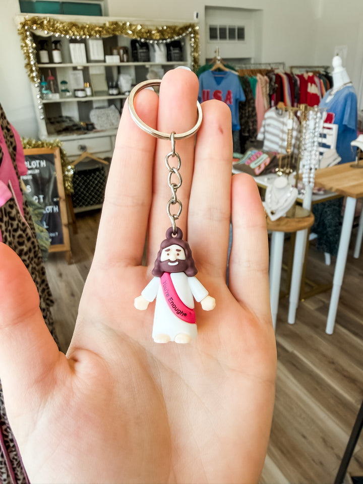 A hand holding a small Jesus figurine keychain with a silver split ring, set against a blurred boutique background.