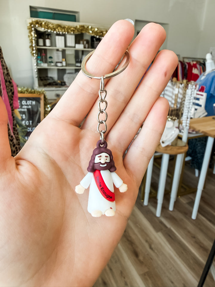 A hand holding a small Jesus figurine keychain with a silver split ring, set against a blurred store background.