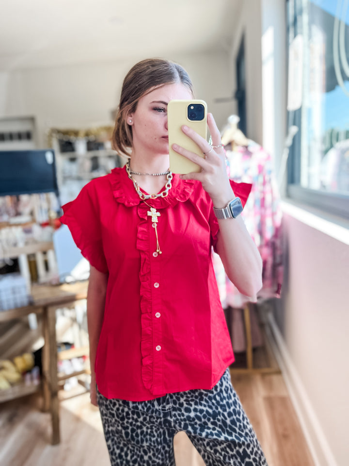 A woman takes a mirror selfie in a boutique, wearing a bright red short-sleeve blouse with ruffles and a gold cross pendant, paired with leopard print pants.