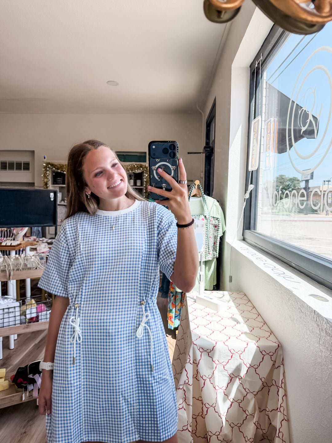 A woman taking a mirror selfie in a boutique shop, wearing a light blue and white gingham short-sleeve dress with drawstrings at the waist.